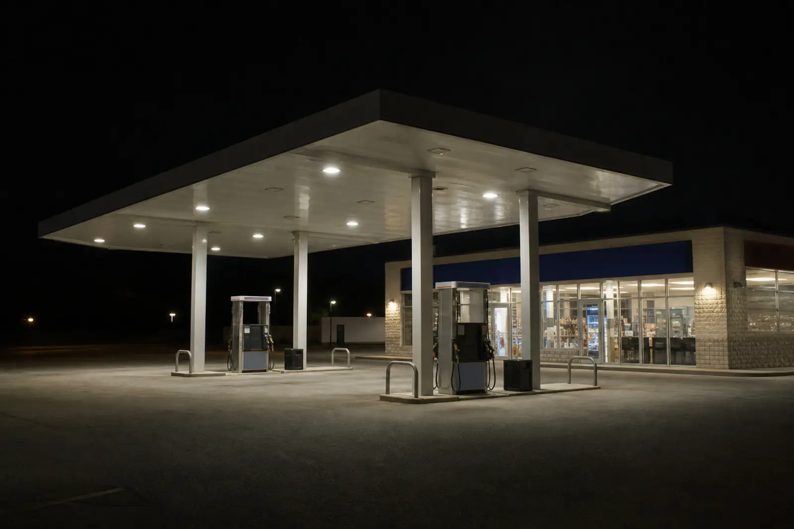 Documentary photograph of a convenience store and fuel canopy at night, unbranded fuel pumps under white canopy lights, empty forecourt, no people, no readable