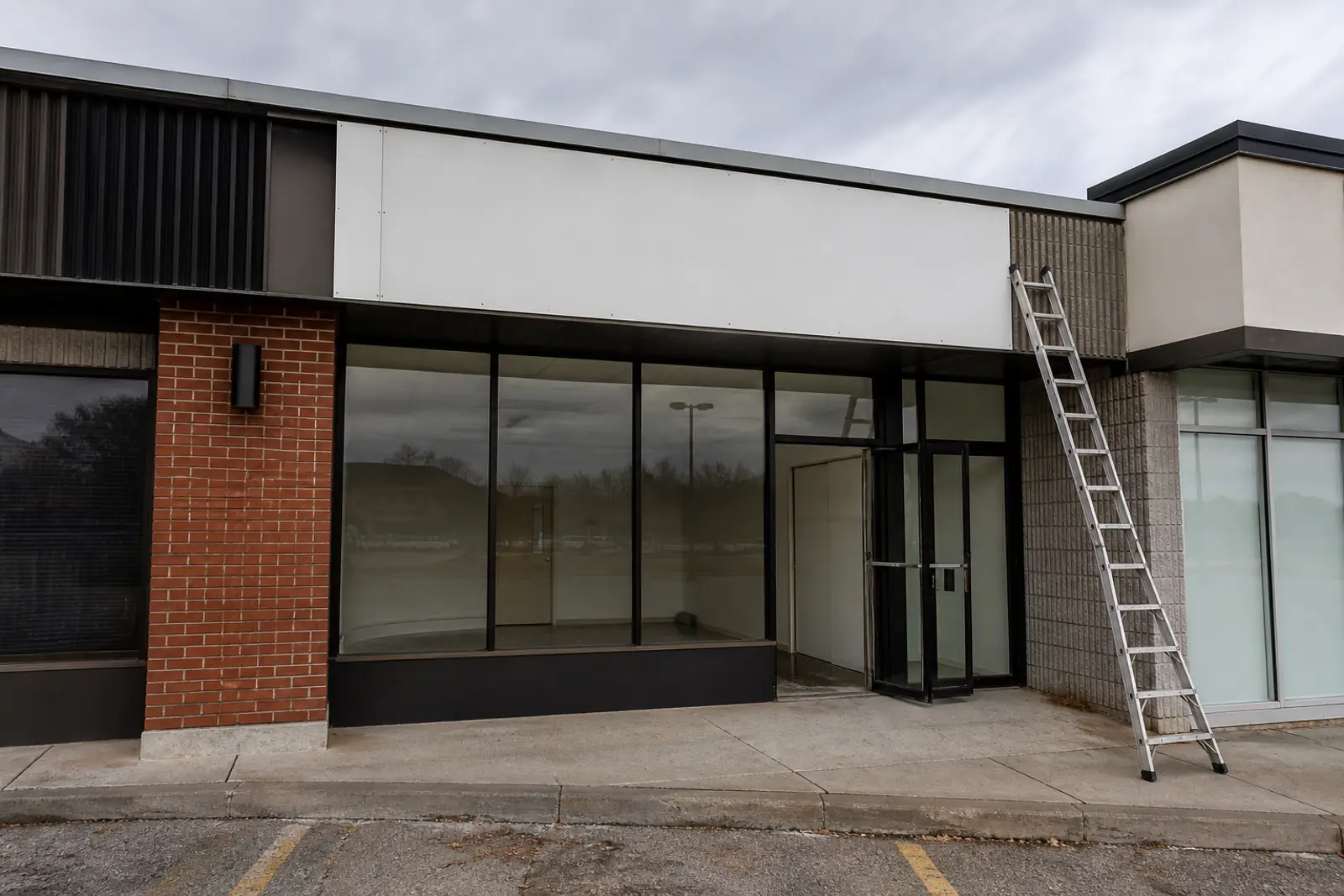 A strip-mall storefront mid-changeover: a blank white sign panel above the door, an aluminum ladder leaning against the facade, empty display window, overcast d
