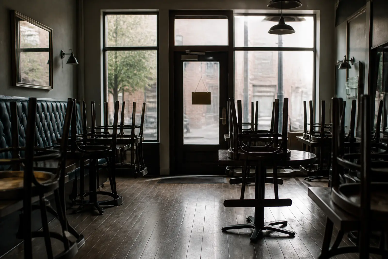 Documentary photograph of a quiet restaurant dining room in 2020: chairs turned up on tables, soft window light, a blank closed sign on the glass door