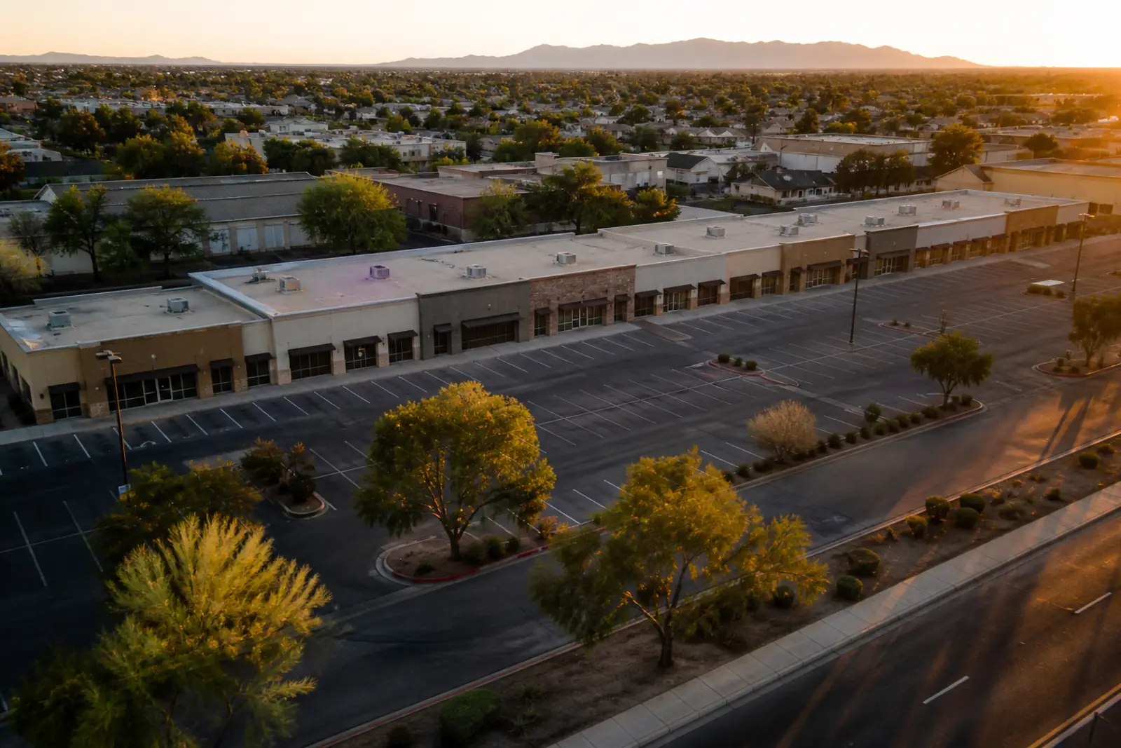 Aerial view of a suburban commercial strip at golden hour, a long row of small single-story storefronts with plain unbranded signs, parking lots and a few trees