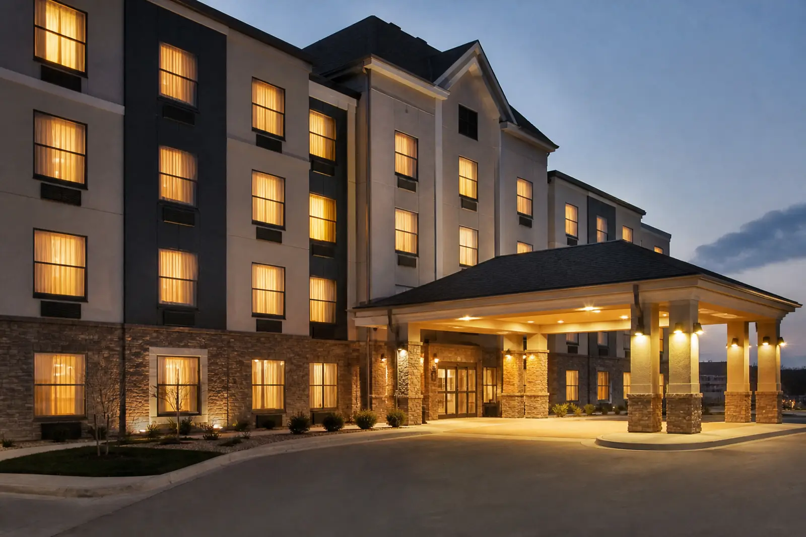Documentary photograph of a mid-size hotel exterior at dusk, rows of lit windows, an empty covered portico with warm light, no people, no readable signs, logos